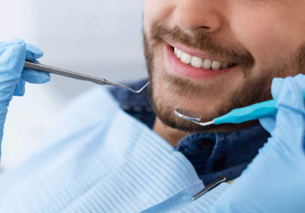 Closeup of dental clinic patient smile and dentist hands holding tools, unrecognizable cheerful bearded man attending dentistry, panorama. Banner for modern dental clinic advertisement