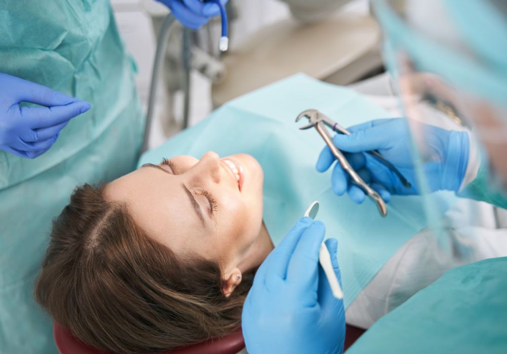 Female patient lying in dental chair and smiling while two dentists standing beside woman and holding dental tools