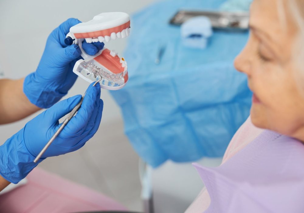 Stomatological specialist operating a dental probe while giving lecture to old woman about tooth implants using mockup